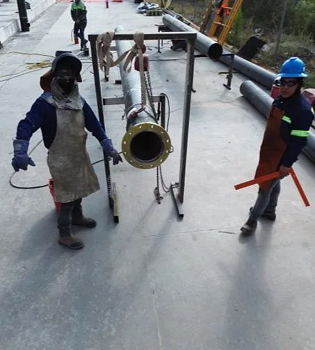 Dos montadores con equipo de protección izando un carrete de tubería bridada